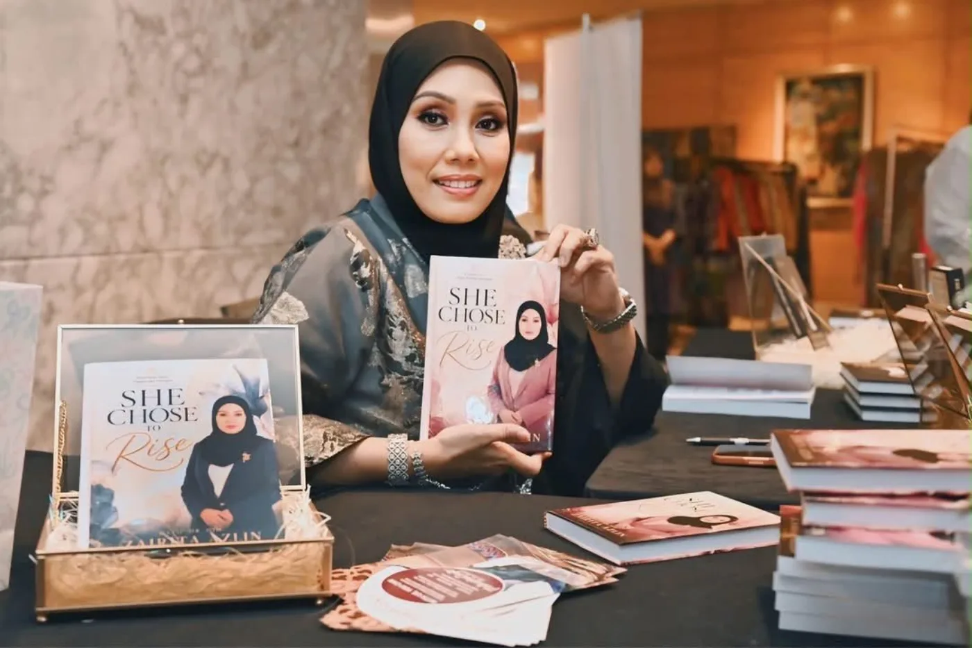 Dato&rsquo; Dr. Hjh. Zairita Azlin holds a copy of She Chose to Rise at the signing table beside stacked copies of the book